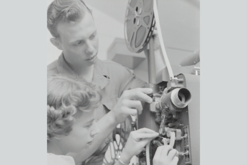 An old photo of a a man working with a woman to set up an old film reel projector.