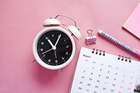 A photo of pink background with an old  fashioned alarm clock with a paper clip, paper calendar, and a pencil.